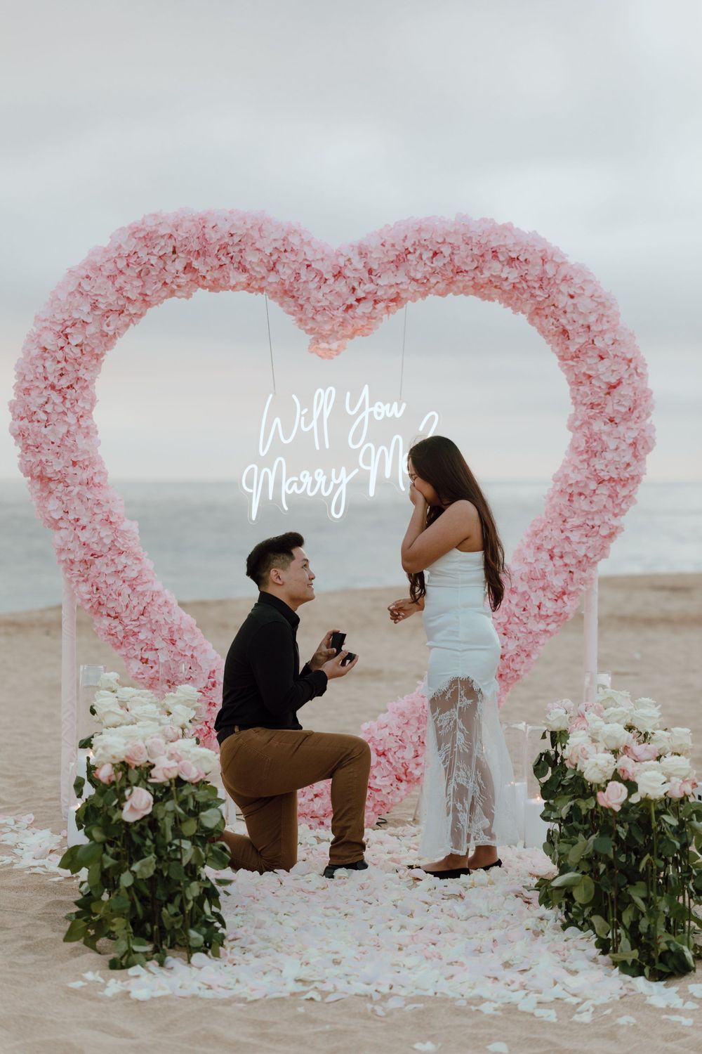 A beach proposal scene with a pink floral heart arch and 'Will You Marry Me' neon sign.