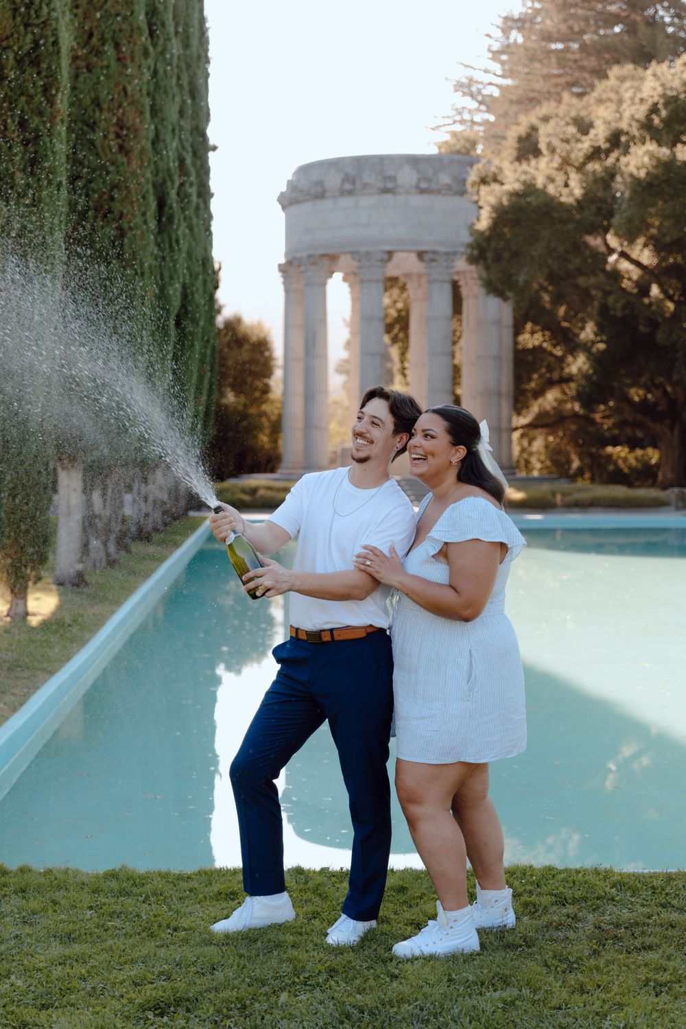 A couple celebrating by spraying champagne near a classical columned structure and pool.
