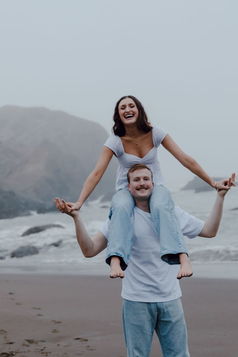 A playful couple enjoys a piggyback ride on a misty beach with mountains in the background.