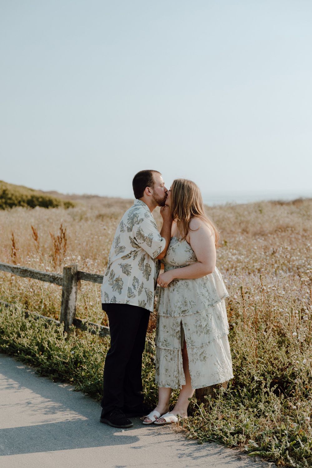 A couple shares a romantic kiss on a coastal path surrounded by golden grass.