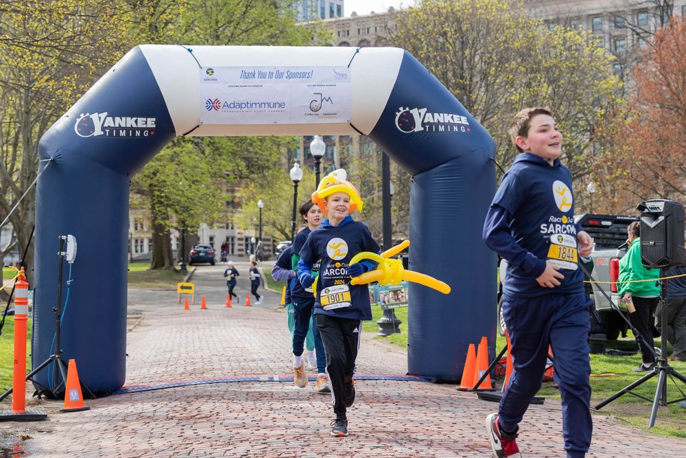 Runners pass under a blue inflatable arch during an outdoor race event on a brick pathway.