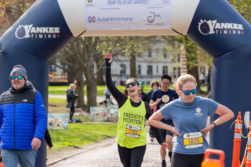 Athletes running through a branded archway during a race event on a tree-lined path.
