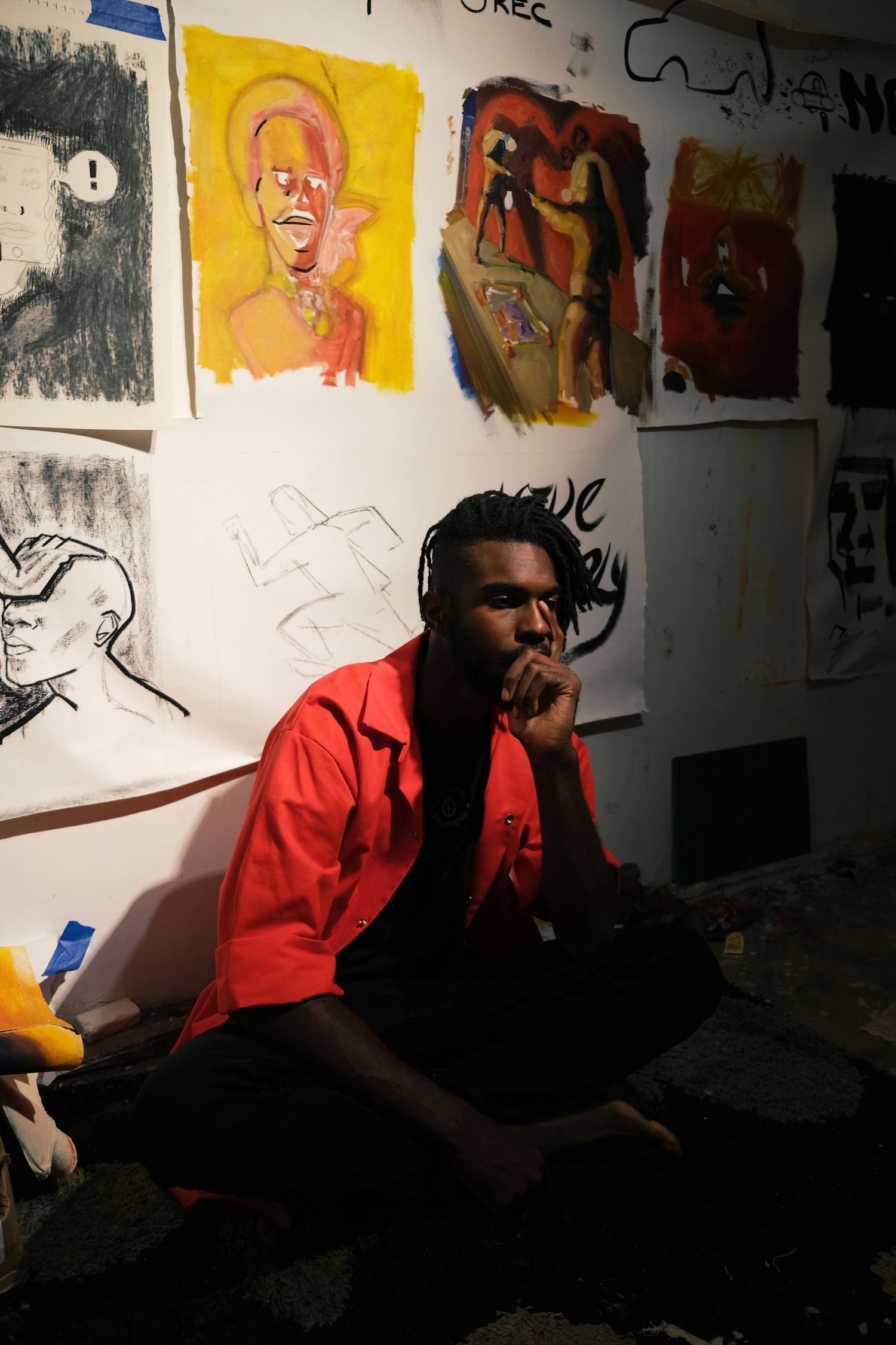 Artistic scene with person in red jacket seated beneath wall of colorful portrait sketches.