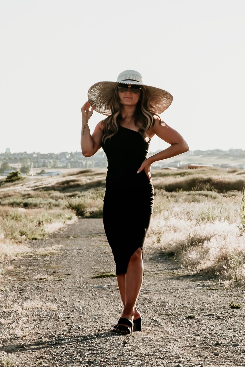 Elegant figure in black dress and wide-brimmed hat poses on dusty trail at sunset.