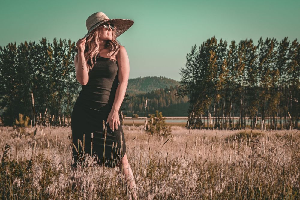 Vintage-toned fashion portrait in black dress and hat among tall grass.