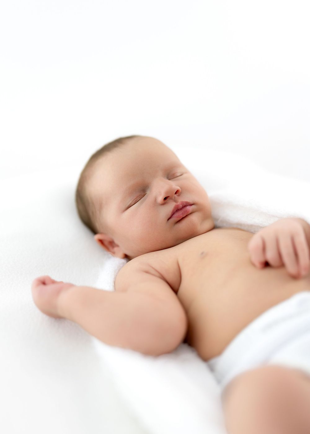 Peaceful sleeping baby lying on white bedding with bare skin visible against soft fabric background.