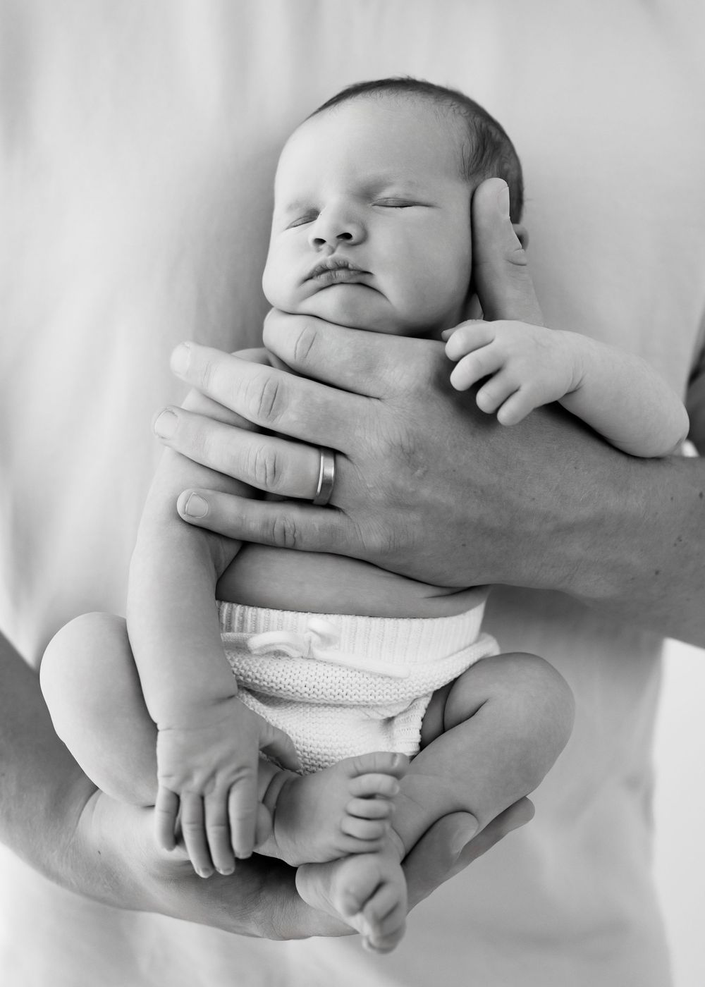 Black and white photo of sleeping newborn baby cradled gently in adult hands while wearing white cloth diaper.