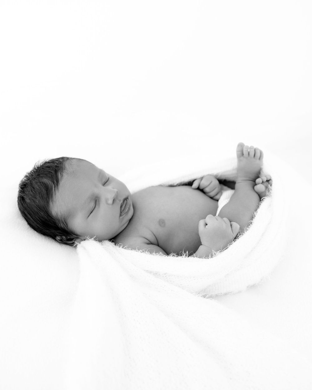 A newborn baby rests peacefully on soft white fabric in black and white tones.
