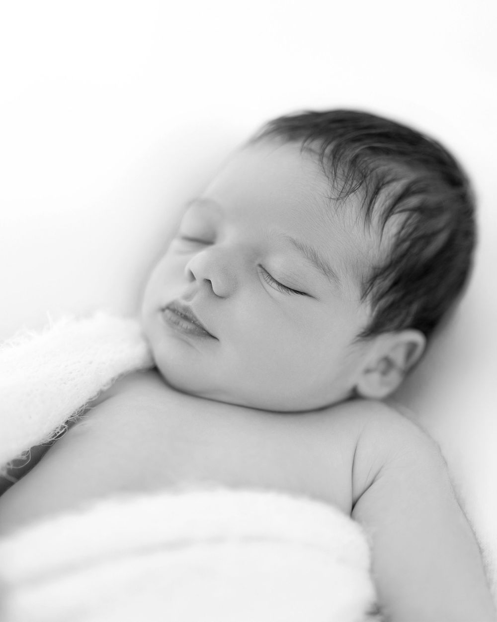 Peaceful sleeping infant wrapped in soft white blanket in black and white portrait.