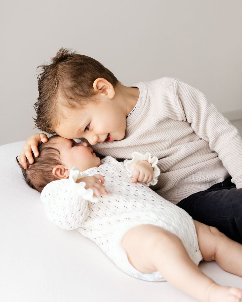 Sibling shows affection to baby wearing white lace outfit while lying together on white surface.