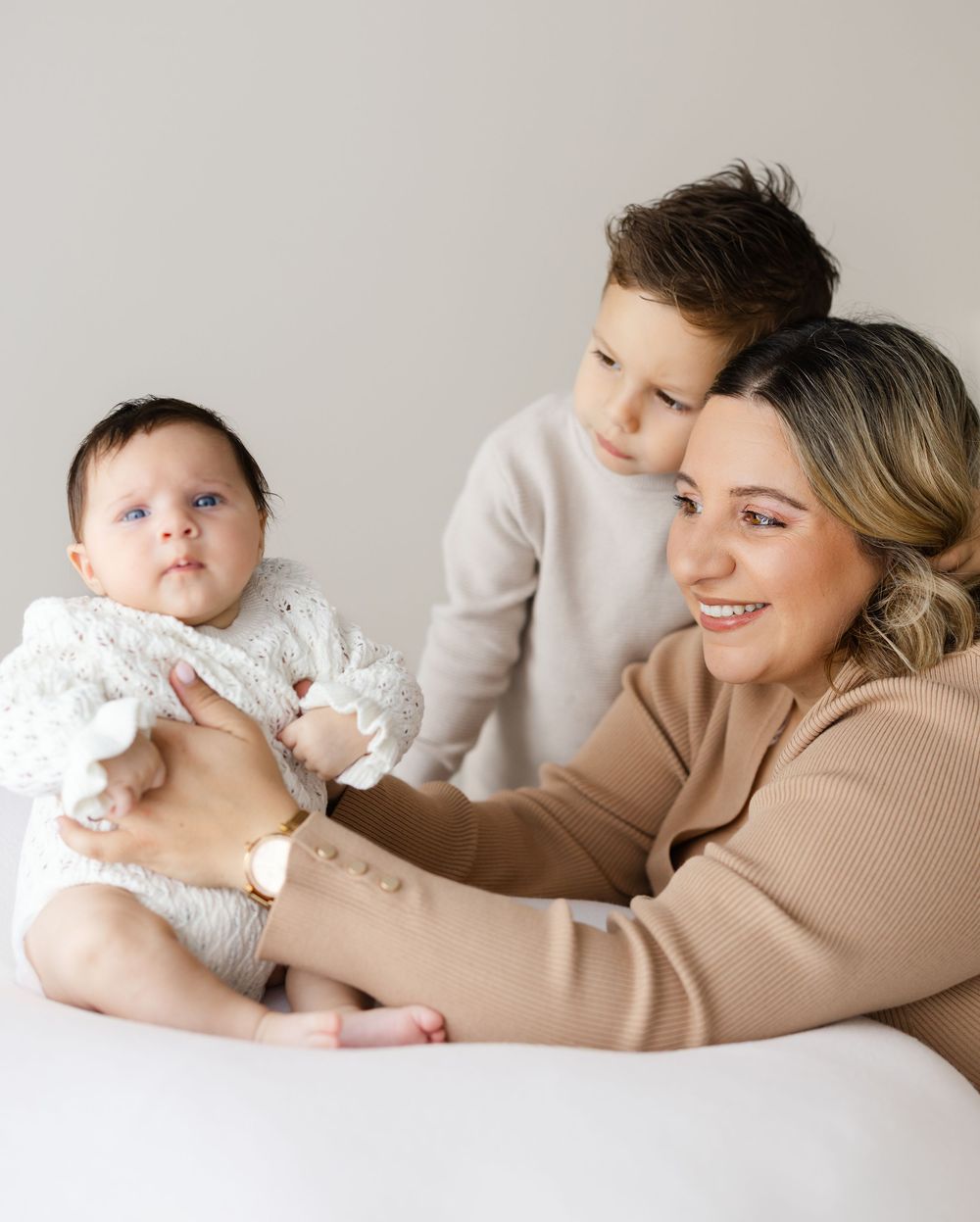 Mother and baby in beige sweater lie together on white backdrop during minimalist photo session.
