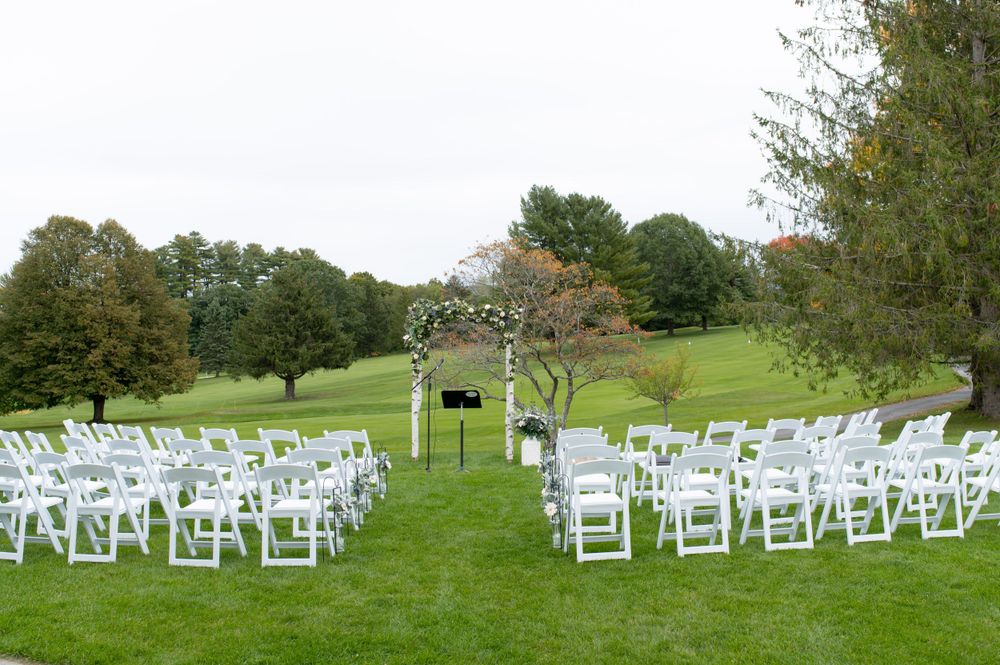 Wedding ceremony setup with white chairs and floral arch on manicured lawn with rolling hills backdrop.