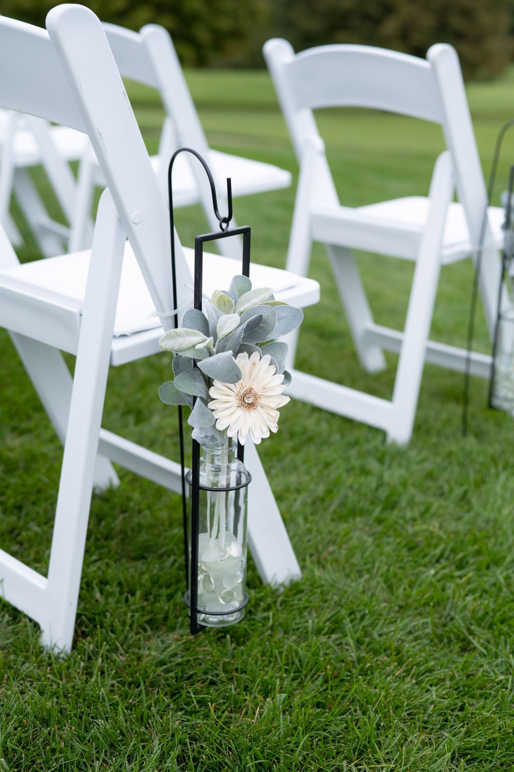 White ceremony chairs decorated with simple flower arrangements in glass vases on green grass.
