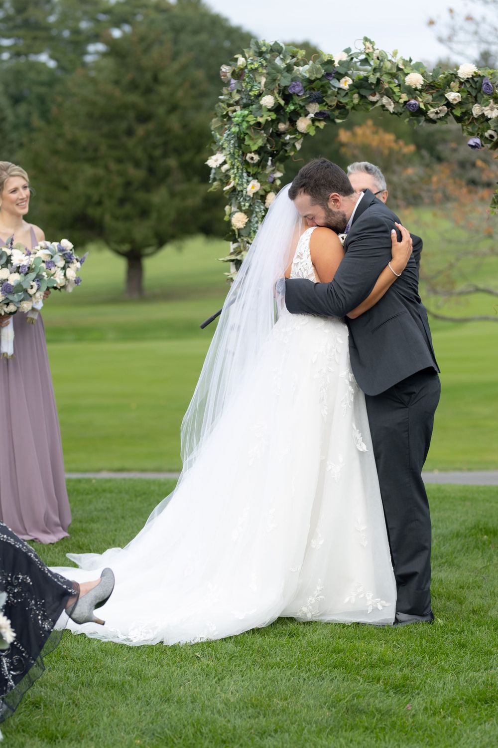 Romantic embrace under floral wedding arch with bridesmaid in purple dress visible.