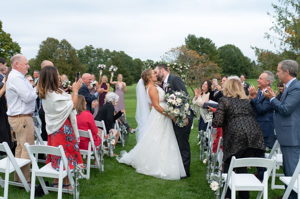 Wedding guests throw flower petals as the newlyweds walk down the aisle on a grassy lawn.