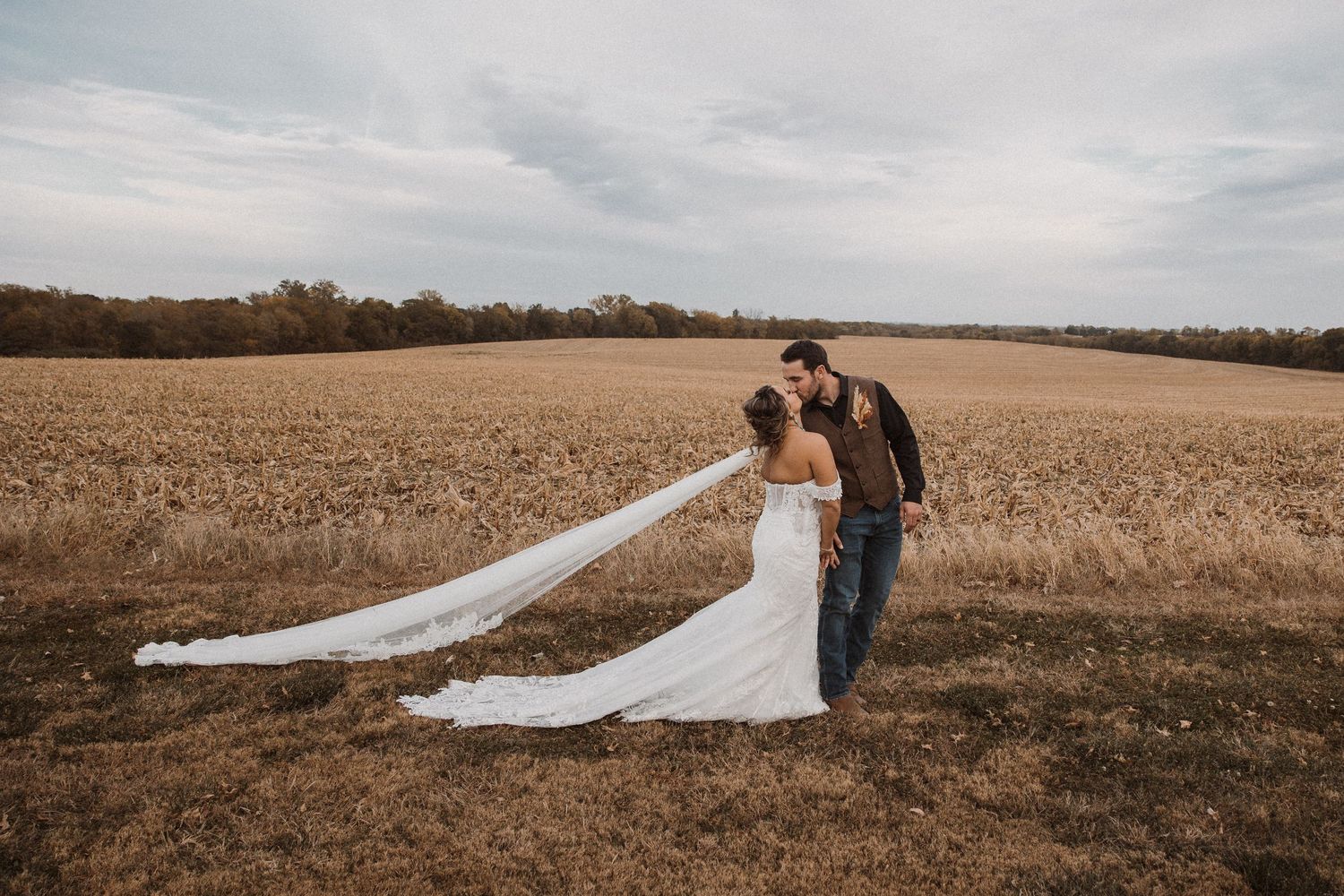 A couple in wedding attire standing in a golden wheat field with flowing veil.