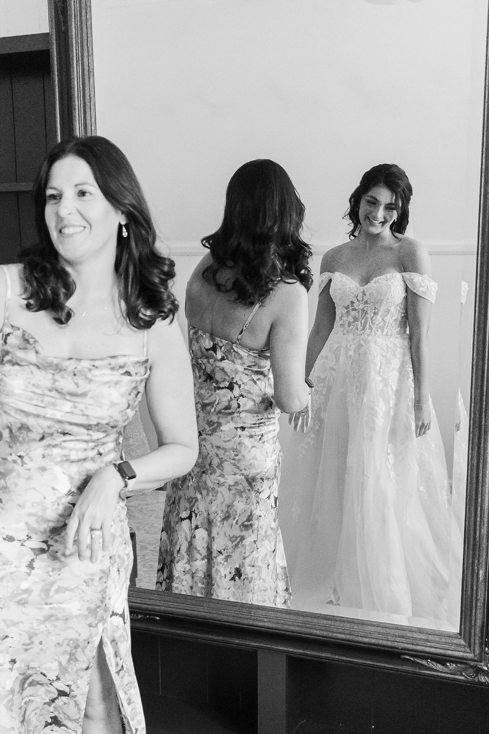 A bridal party gets ready in a room with a large mirror reflecting their elegant dresses.