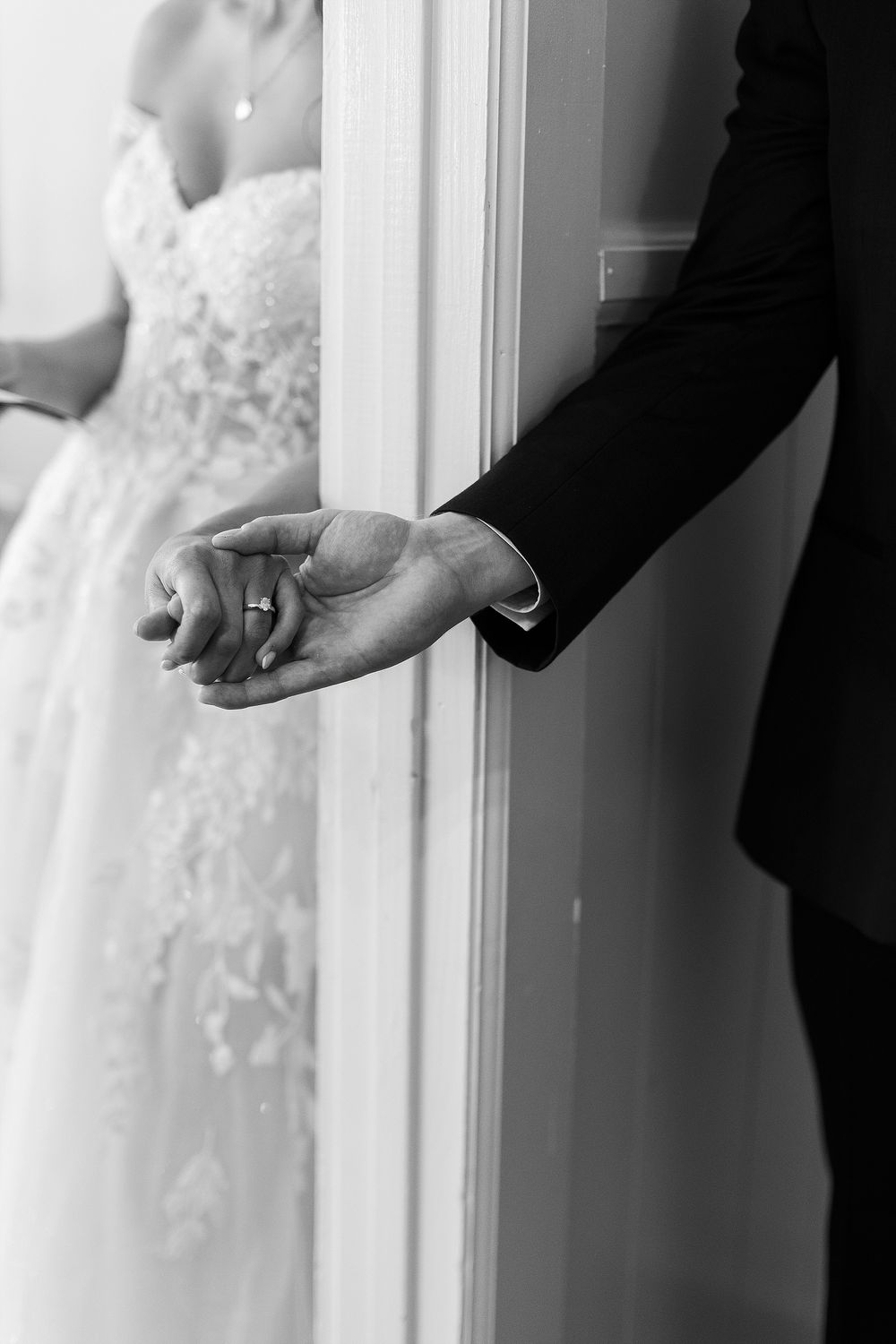 Black and white photo of couple holding hands through doorway, bride in white dress.