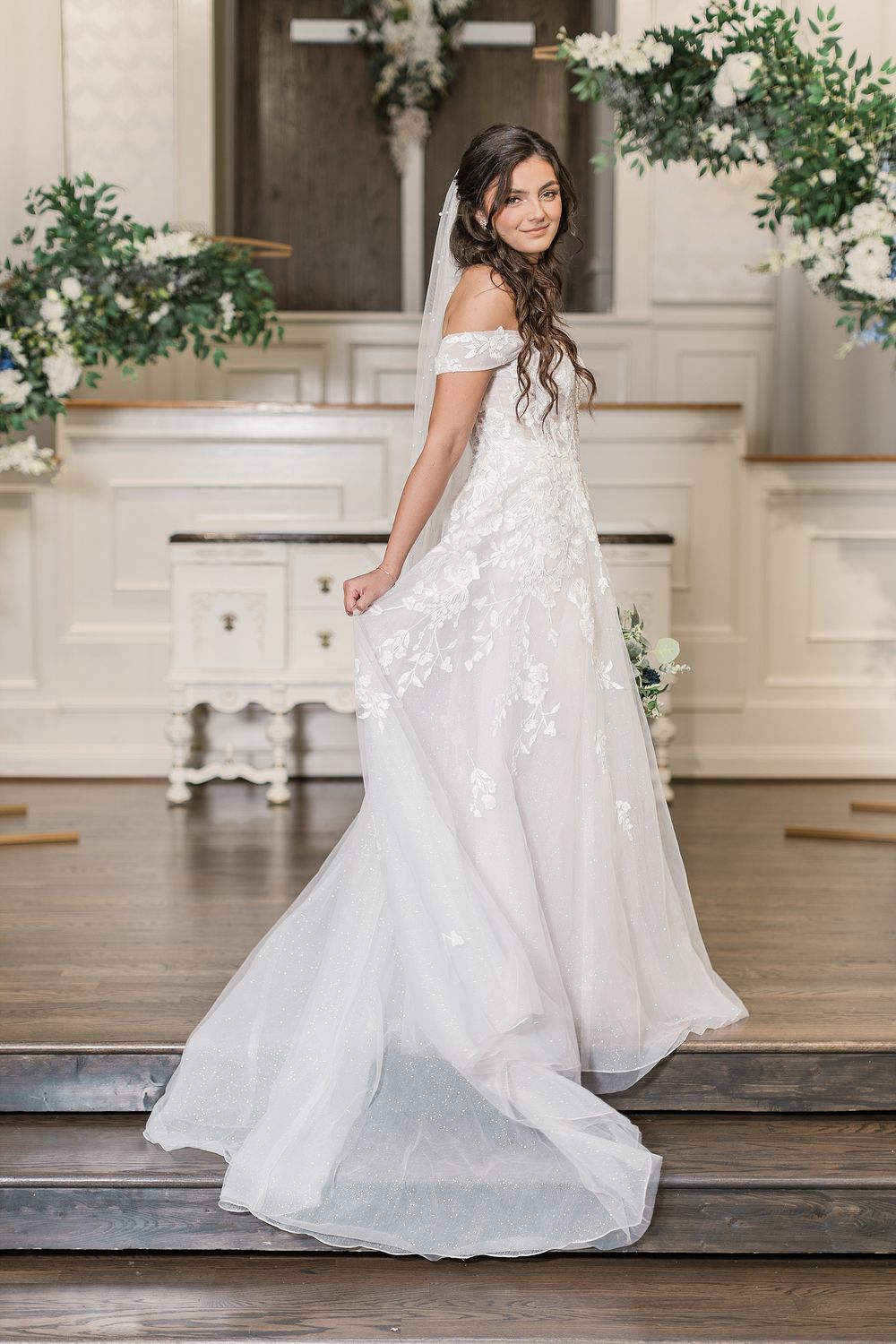 Bride in off-shoulder lace gown poses on church steps with floral decorations.