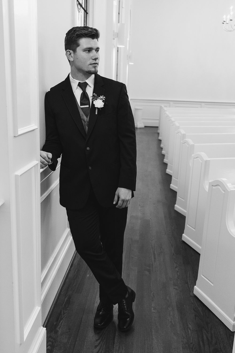 Groom in classic black suit stands in church hallway with white pews.