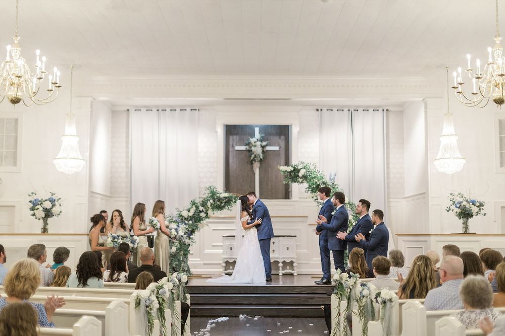 Wide view of wedding ceremony in traditional white church with guests seated in wooden pews.