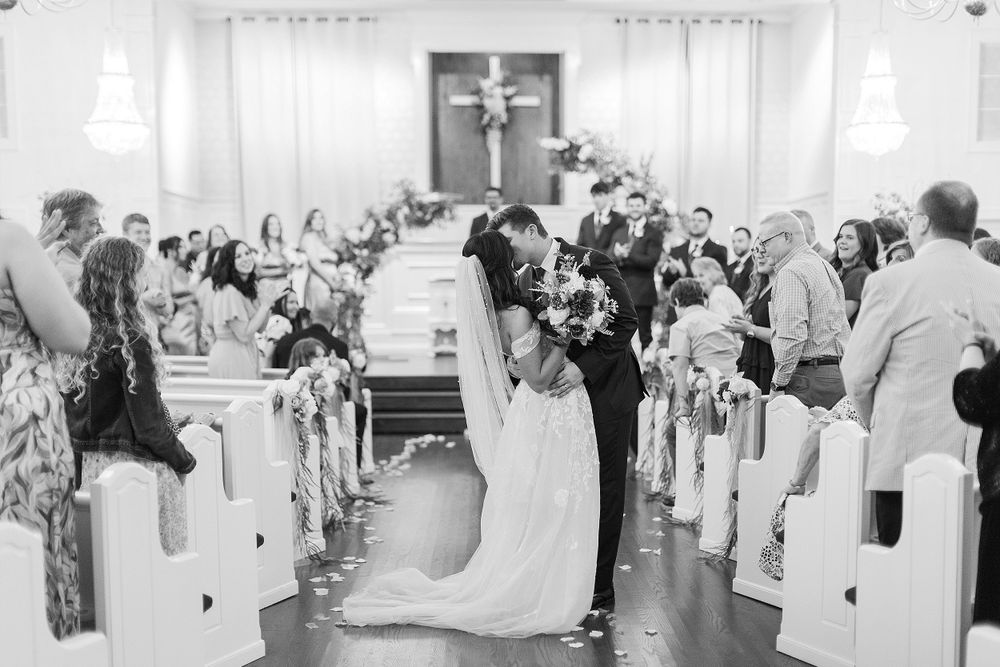Black and white photo of wedding ceremony recessional in church aisle with guests standing.