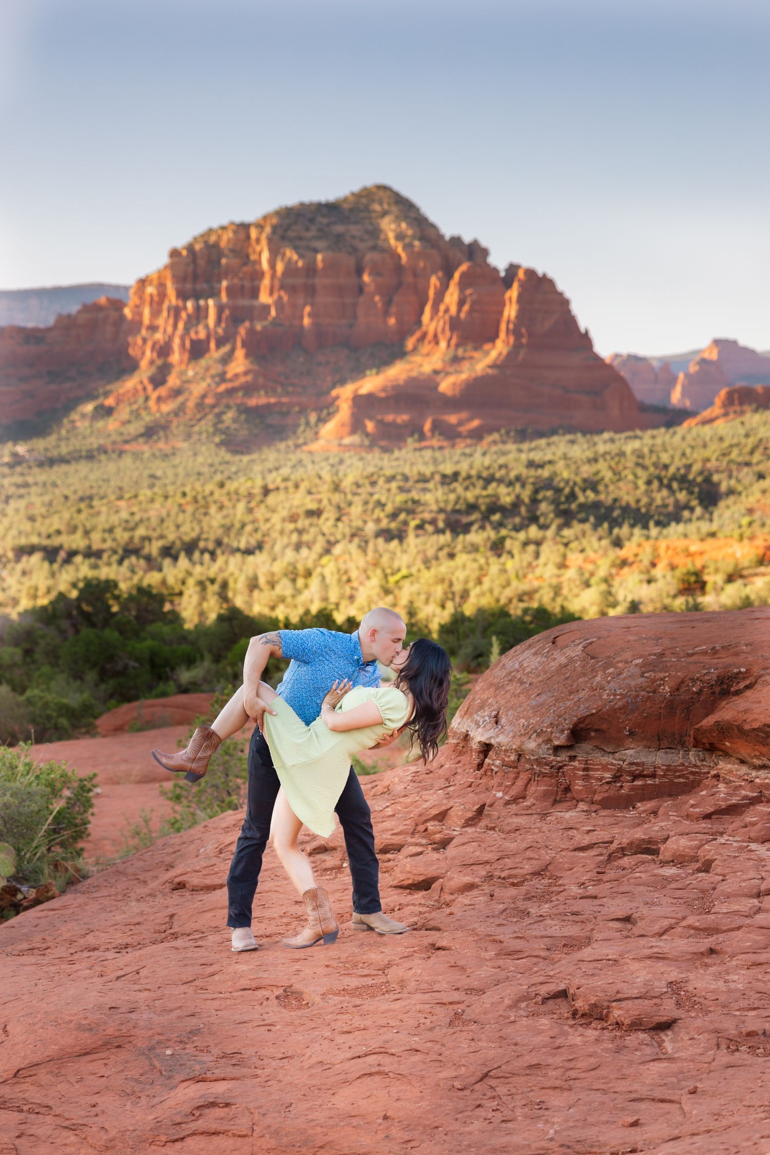 A couple embraces on red rocks with majestic Sedona buttes and desert landscape in the background.