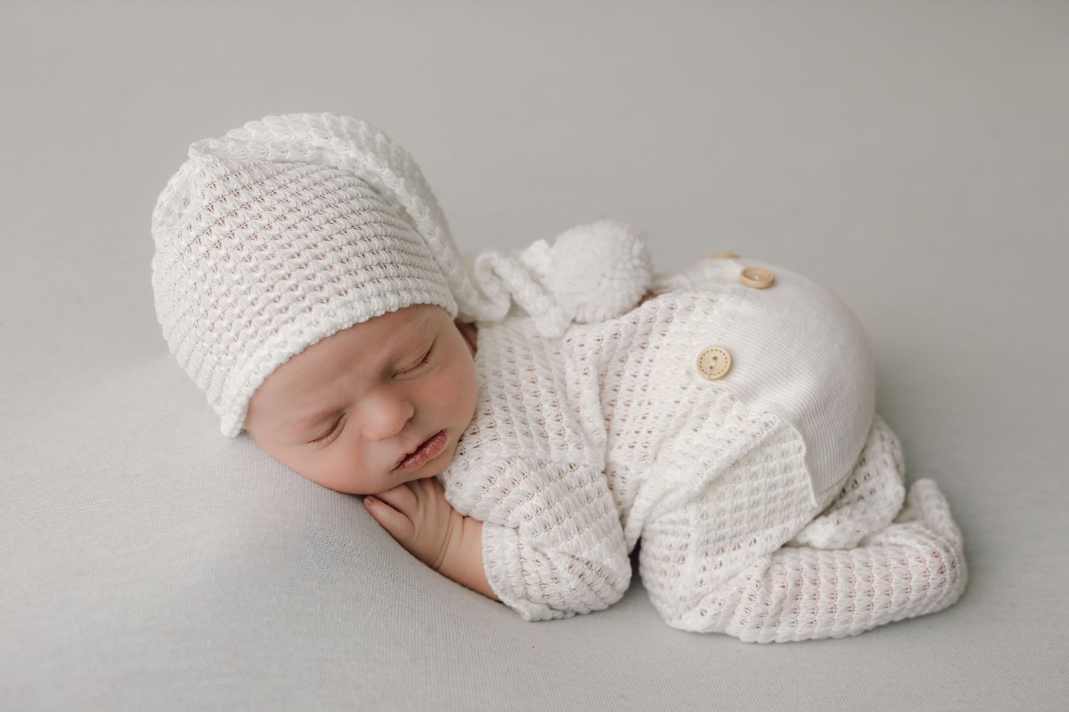 Baby dressed in a white newborn outfit on a plain white backdrop blanket posed laying on his tummy for his studio newborn photo session.