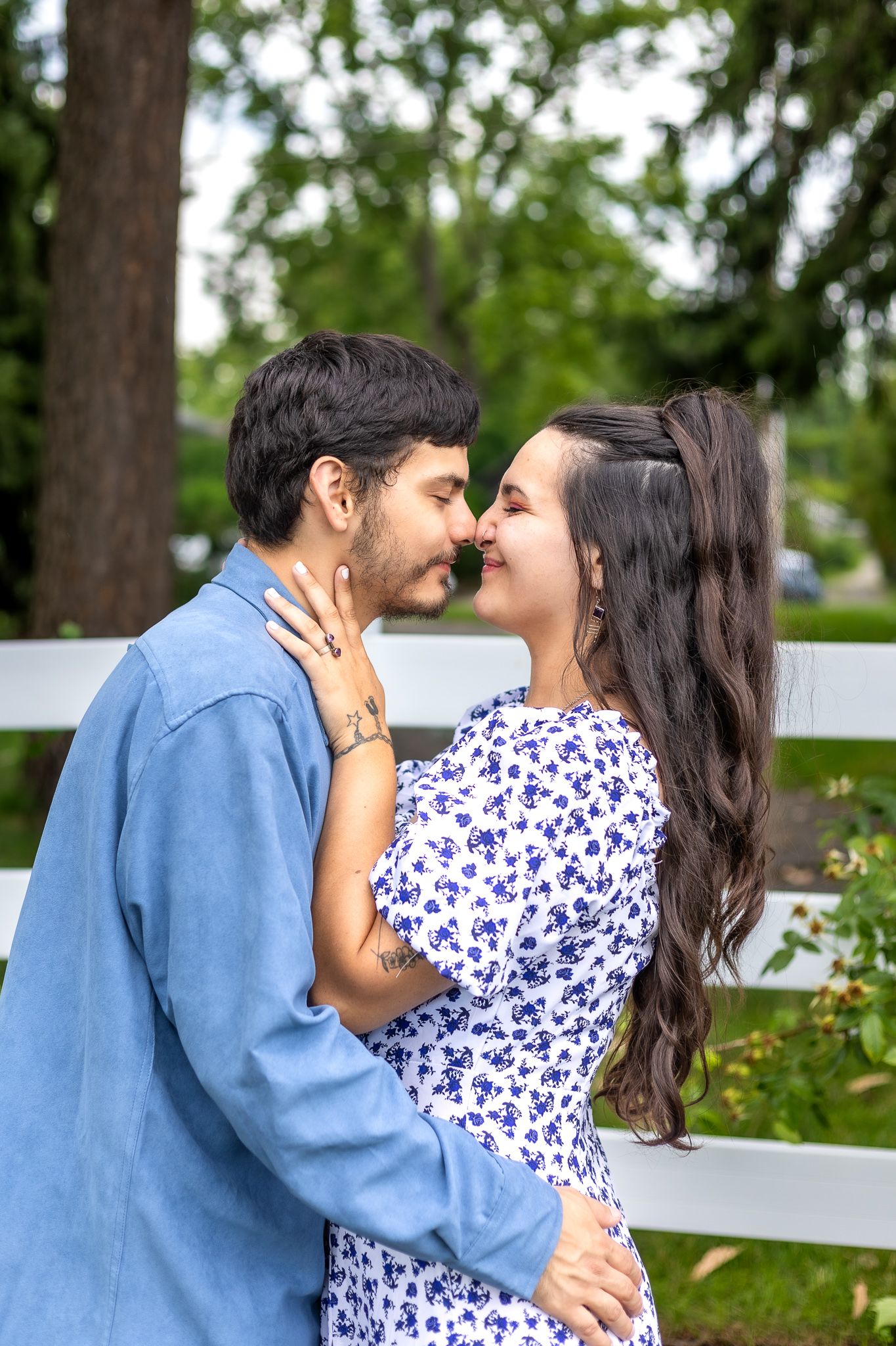 A romantic couple embraces in front of a white picket fence on a summer day.