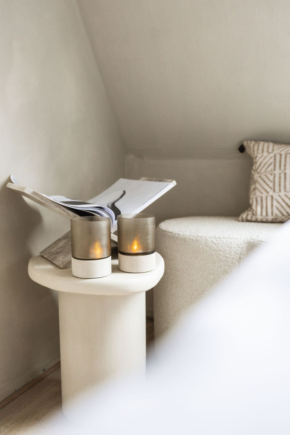 A minimalist white side table displays two lit candles and a book next to a textured throw pillow in a cozy corner.