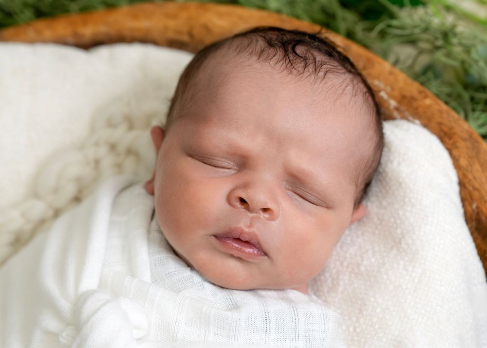 Peaceful newborn in white clothing sleeping in a natural wood bowl setting.
