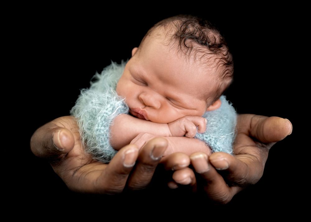 Sleeping newborn in soft blue wrap gently cradled in hands against dark background.