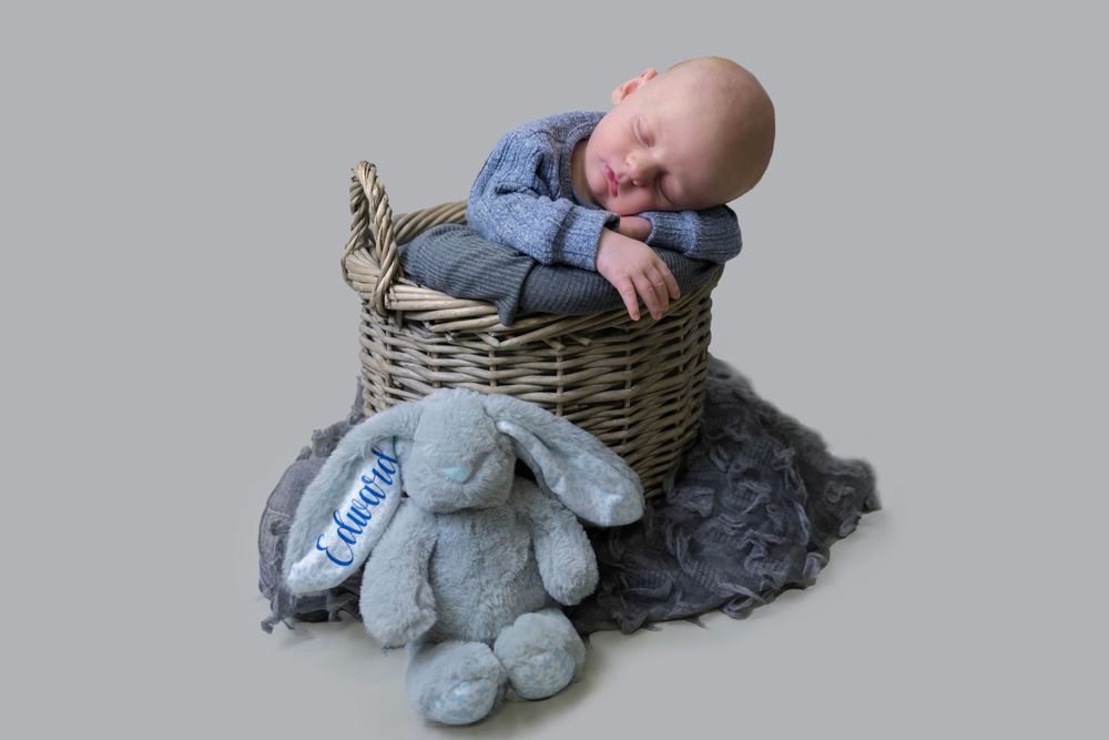 Sleeping baby in blue outfit resting in wicker basket with plush toy.
