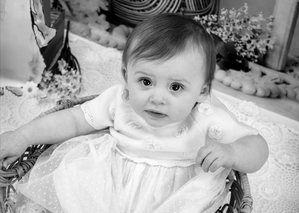 Black and white portrait of baby in white dress sitting on lace.