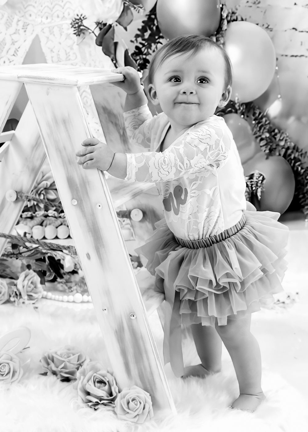 Black and white photo of baby in tutu dress standing by decorative ladder with balloons.