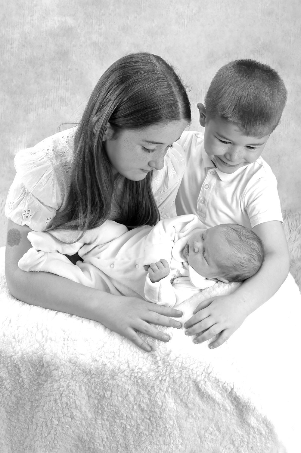 Black and white image of siblings looking tenderly at their newborn family member.