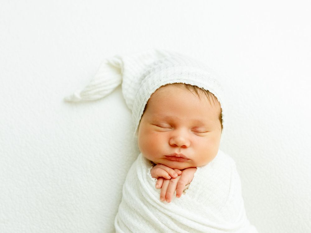 A sleeping newborn baby wrapped in white fabric wearing a knitted white hat with a pompom on top.