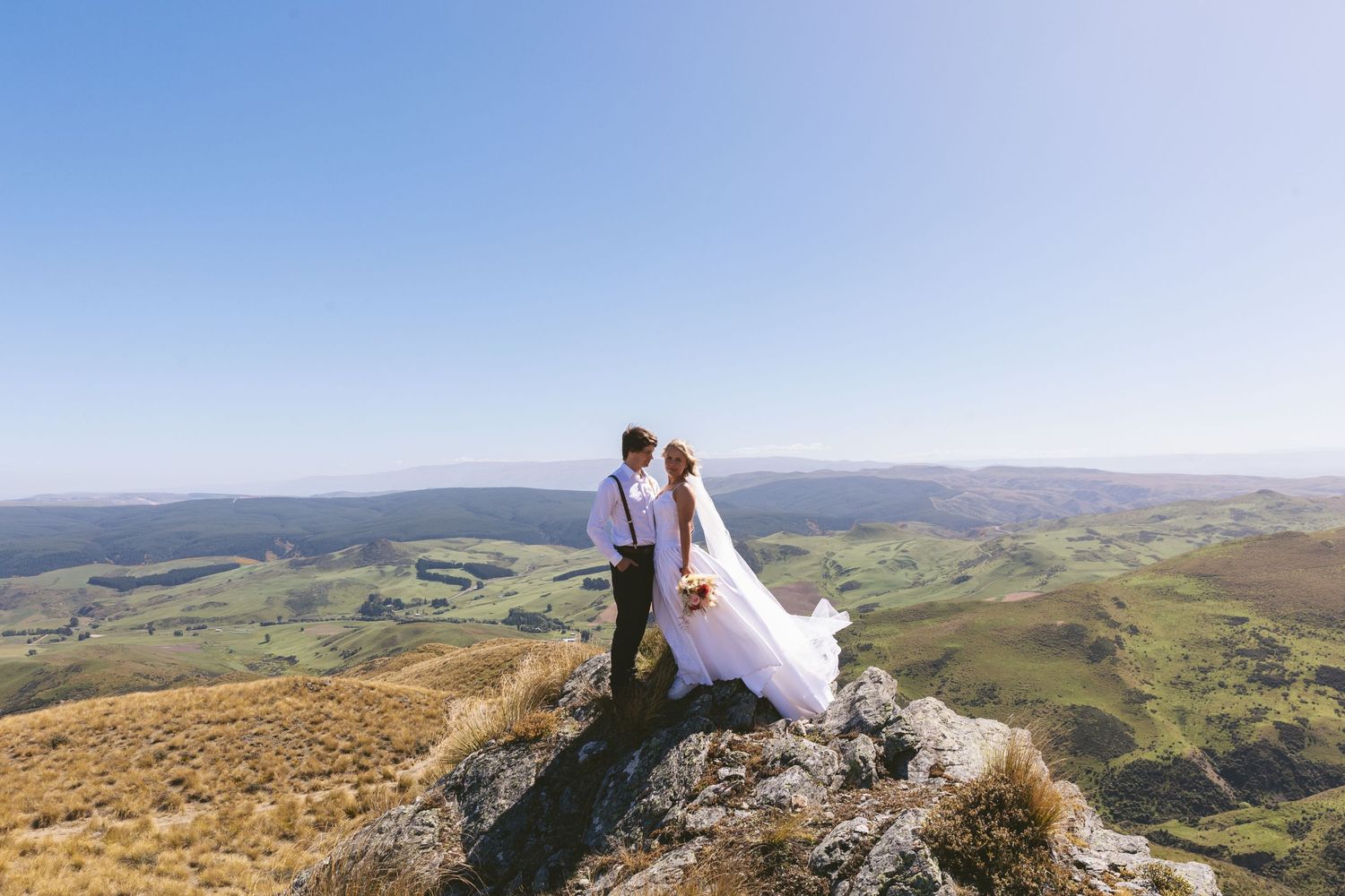 Newlyweds stand together on a rocky mountain peak overlooking a vast valley landscape.