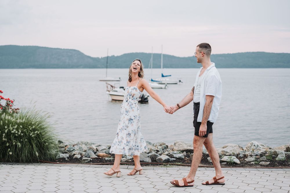 An engaged couple hold hands while walking along a Hudson River path with boats and hills visible in the background.