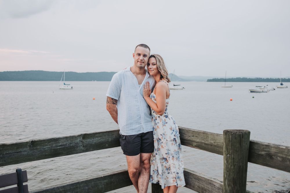 An engaged couple stand on a dock overlooking the Hudson River with boats in the background.