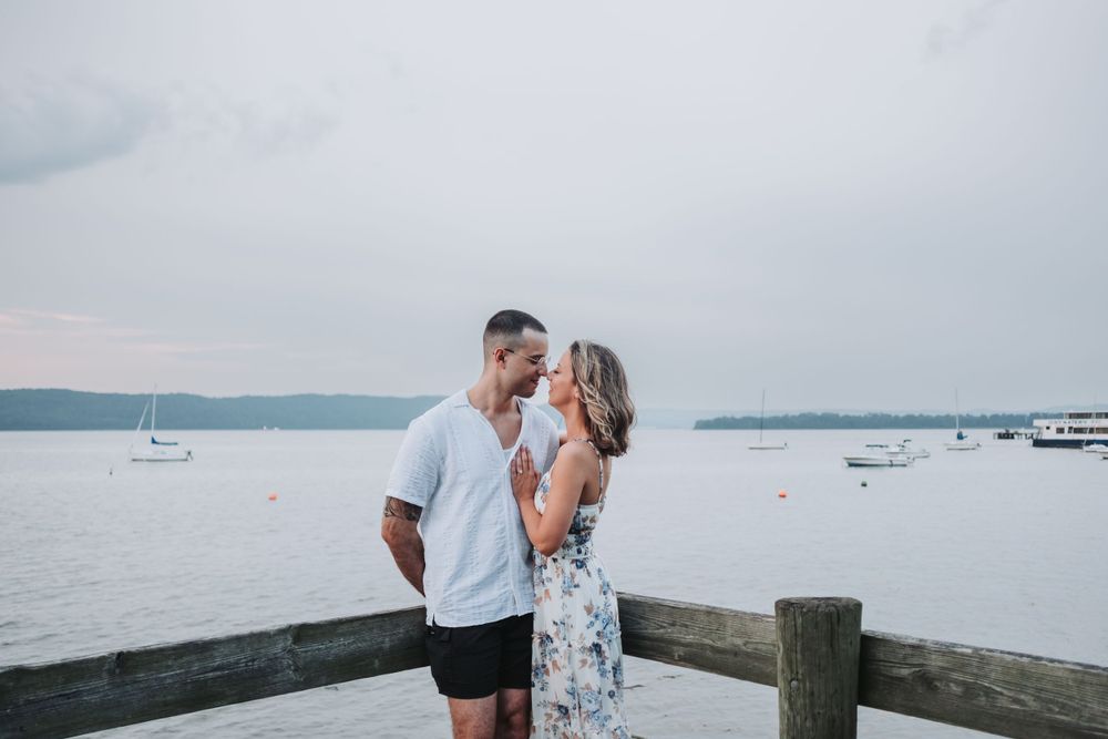An engaged couple stand on a dock overlooking the Hudson River with boats in the background.
