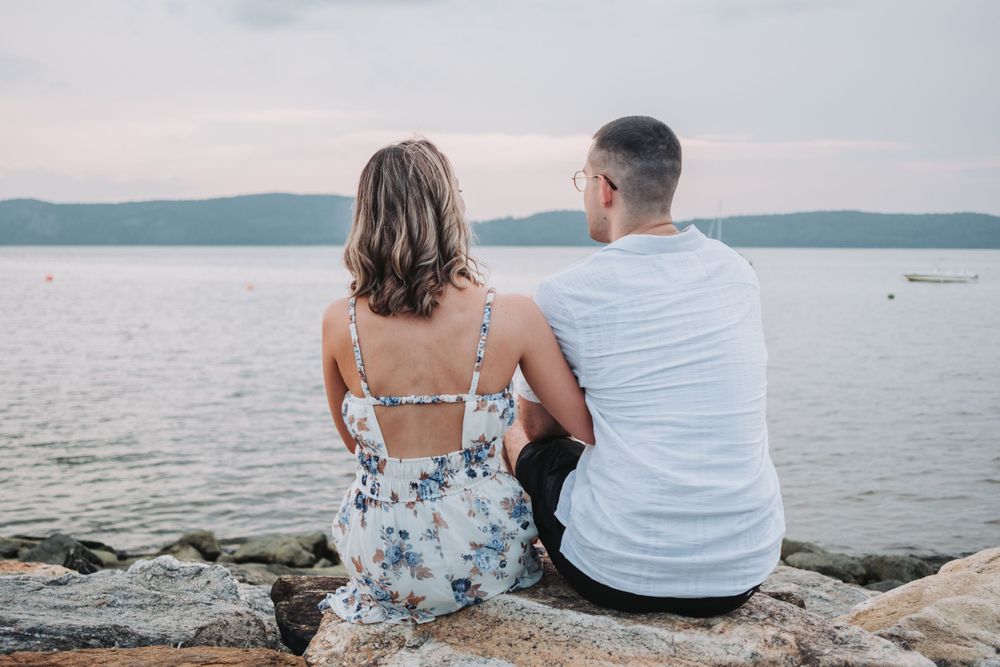 An engaged couple sits together on rocky shore overlooking the tranquil Hudson River at dusk in floral dress and white shirt.