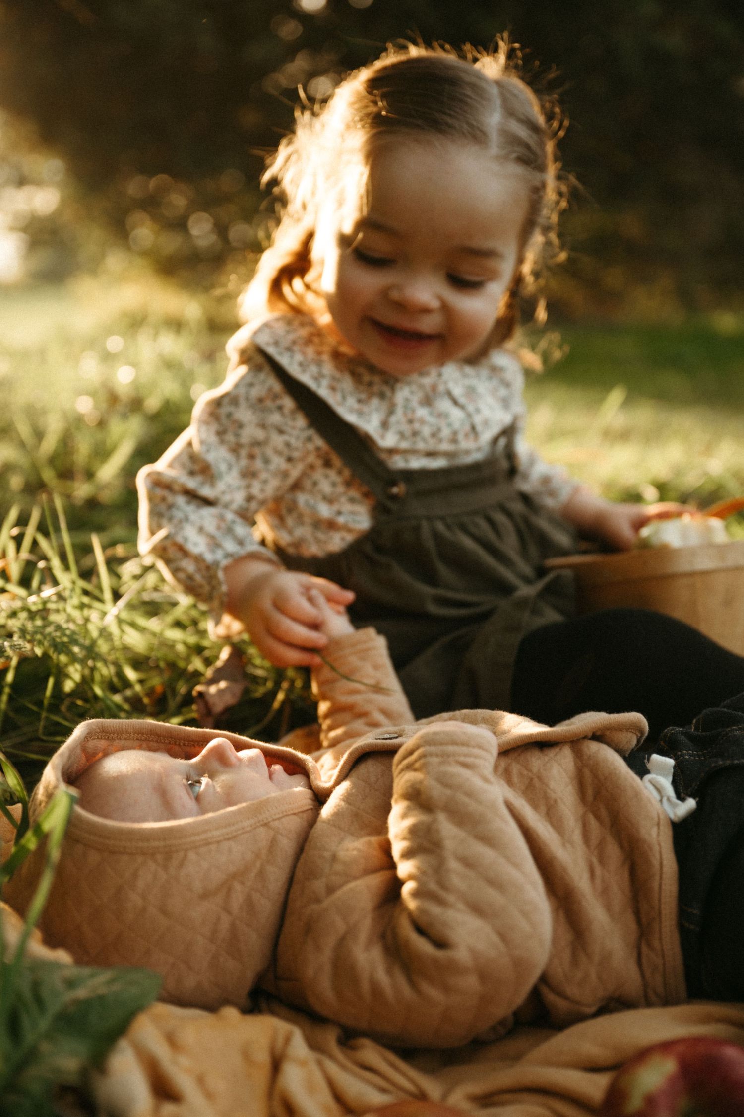 A child in a floral dress sits in sunlit grass beside a brown blanket during a warm autumn afternoon.