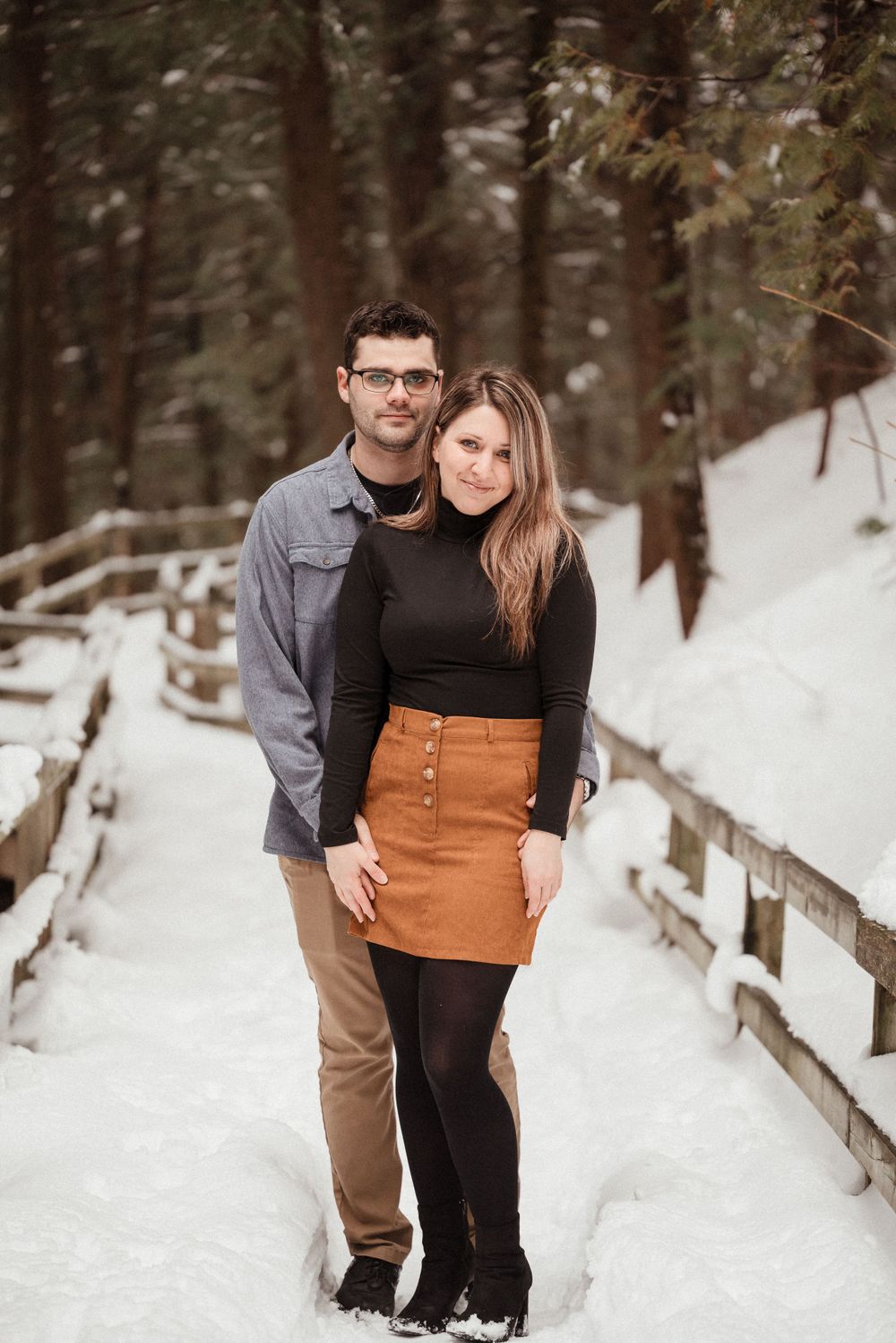 A couple poses together on a snowy wooden bridge surrounded by forest trees and winter scenery.
