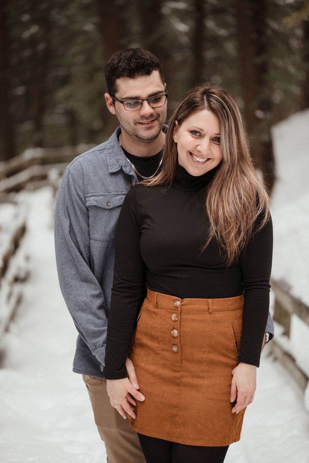 A romantic winter portrait of a couple standing close on a snowy trail in the woods.