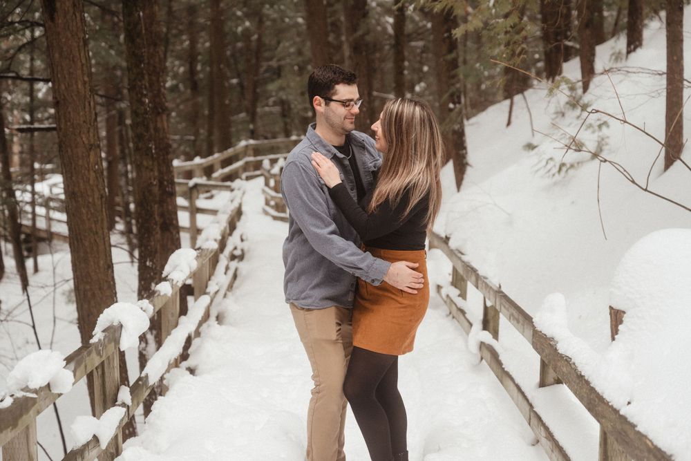 A couple embraces while standing on a wooden bridge path surrounded by snowy forest.