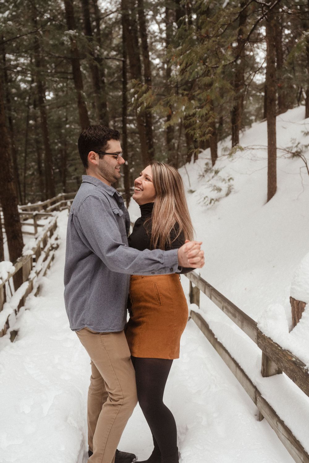 A couple shares an intimate moment while standing on a snow-covered forest walkway.