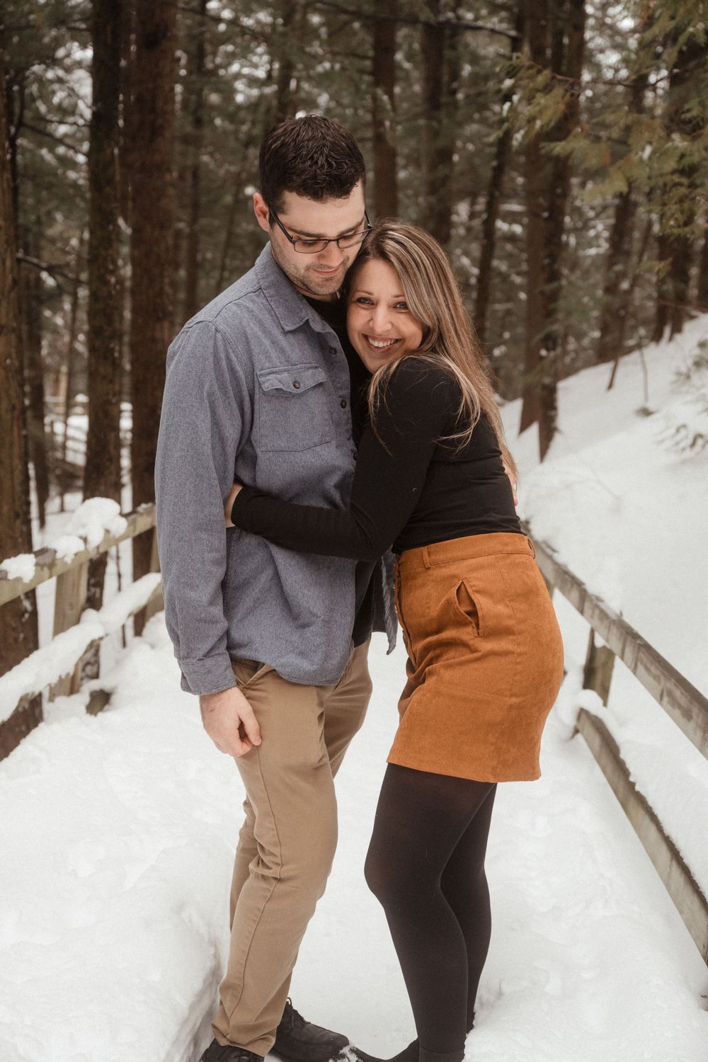A couple embraces while standing on a snow-covered wooden walkway in a peaceful forest setting.