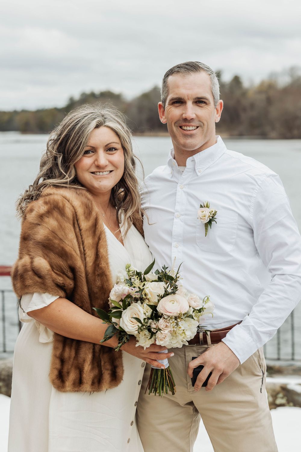 A newlywed couple poses with a bouquet by a lake, the bride wearing a fur wrap.
