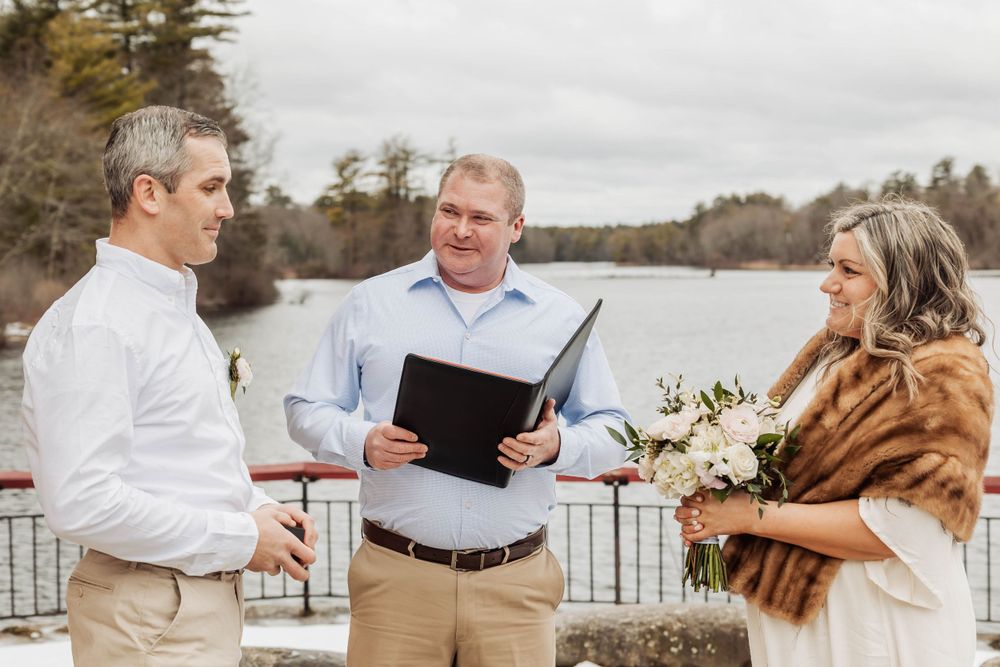 An intimate wedding ceremony takes place by a lake with trees in the background.